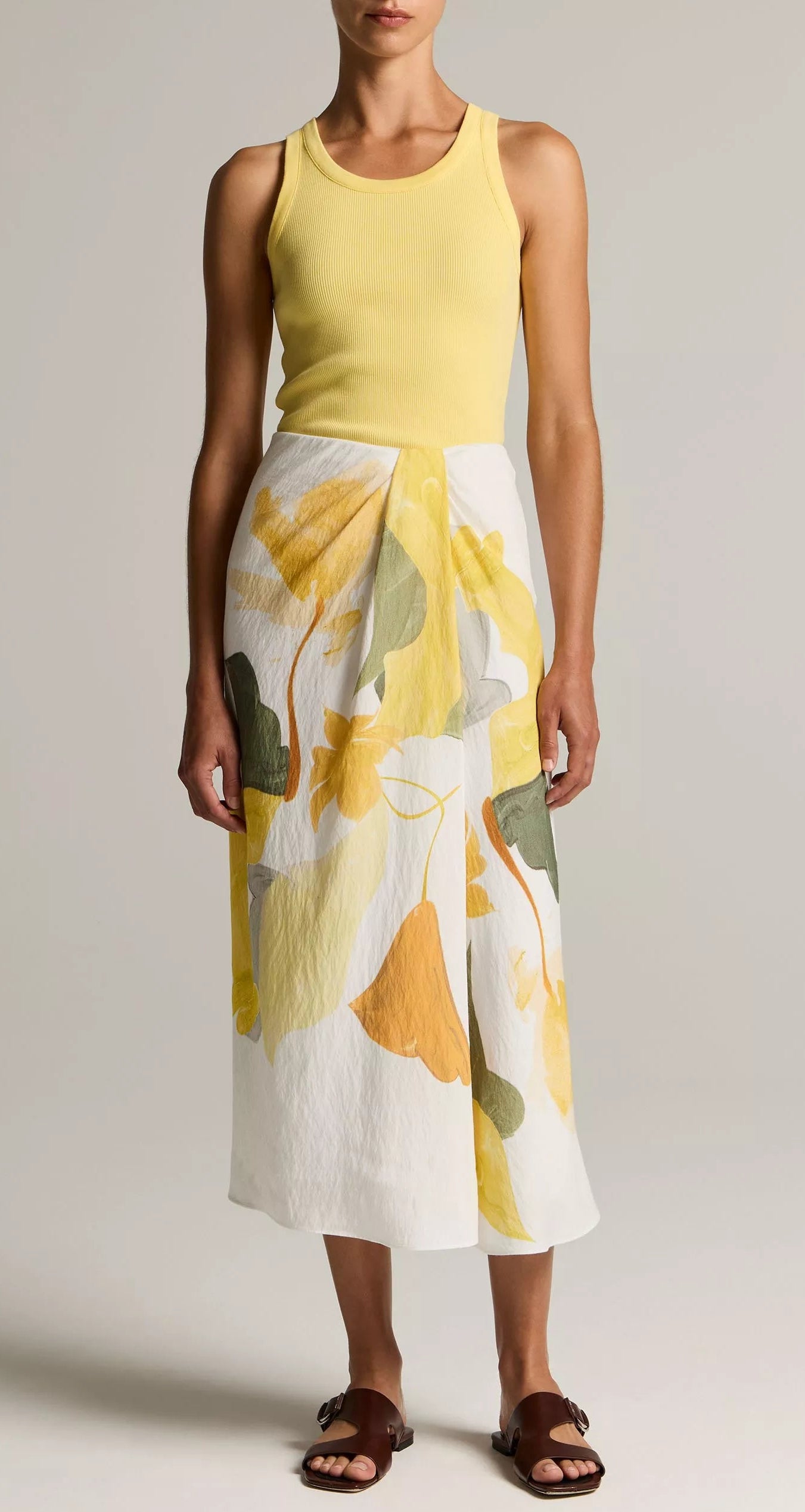 A woman wearing the Vince Ribbed Scoop Neck Tank in yellow, paired with a white midi skirt featuring yellow and green leaf prints and brown sandals, stands against a neutral background.