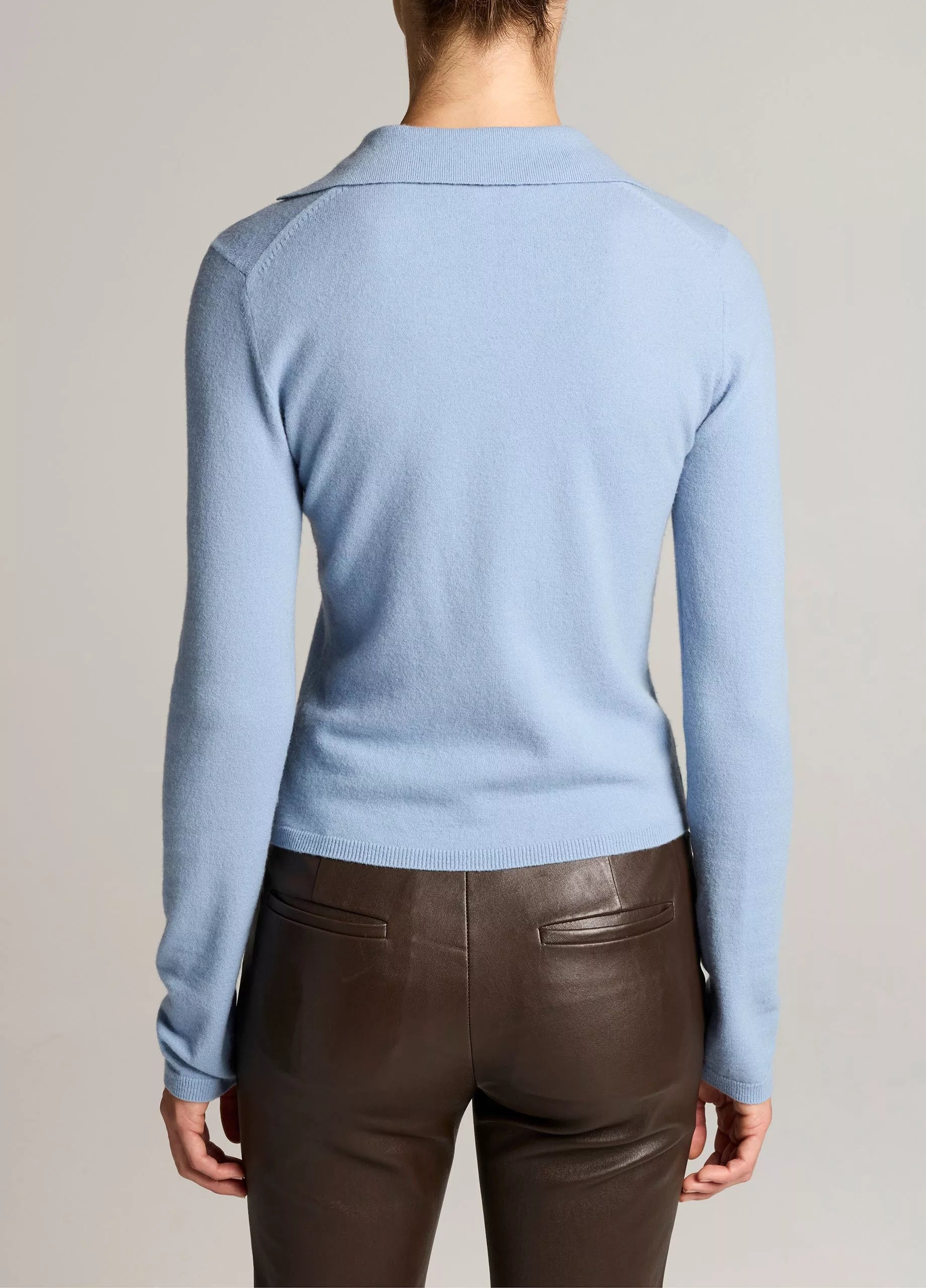 A person is shown from the back against a neutral background, wearing the Vince Long Sleeve High V-Neck Polo Sweater in light blue and dark brown leather pants.