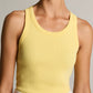 A woman wears the Ribbed Scoop Neck Tank by vince—a sleeveless, finely ribbed yellow top made from a soft cotton modal blend—paired with a white skirt featuring yellow patterns, as she stands against a plain background.
