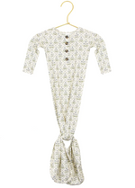 The Lou Lou & Company Baby Girls’ Etta Knotted Gown features long sleeves, a leafy pattern, a buttoned neckline, and a knotted bottom for easy diaper changes, shown on a gold hanger against a white background.