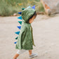 A child in a Great Pretenders Dragon Cape with a soft crushed velvet finish and blue spikes walks outdoors on a sandy path, their imagination taking flight as they transform into a magical creature.