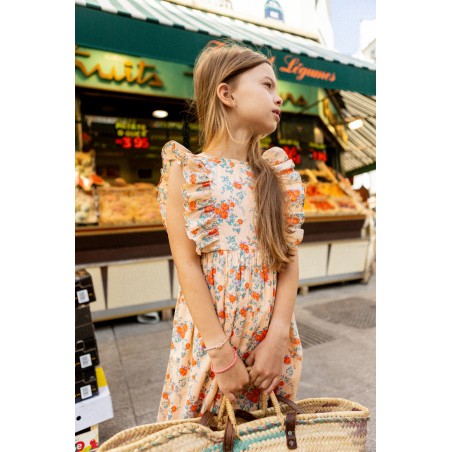 A young girl wearing the Louis Louise Girls’ Arlette Dress holds a woven bag while standing in front of a fruit market stall outdoors.
