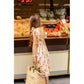A young girl wearing the Louis Louise Girls’ Arlette Dress and carrying a woven bag stands in front of a market produce stand displaying tomatoes, bread, and packaged greens.