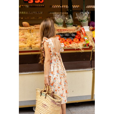 A young girl wearing the Louis Louise Girls’ Arlette Dress and carrying a woven bag stands in front of a market produce stand displaying tomatoes, bread, and packaged greens.
