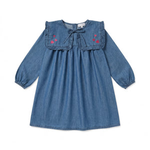 The Girls’ Celicia Dress by Louis Louise is a long-sleeve blue denim dress with a ruffled yoke and square bib collar, featuring a front tie and pink heart embroidery on the chest in a charming flared fit, displayed on a white background.