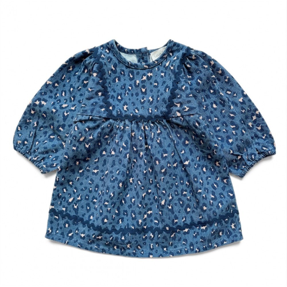 The Baby Girls’ Maha Dress by Louis Louise is a blue, long-sleeve toddler denim dress featuring an all-over leopard print and a gathered flared waist, displayed laid flat on a white background.