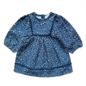 The Baby Girls’ Maha Dress by Louis Louise is a blue, long-sleeve toddler denim dress featuring an all-over leopard print and a gathered flared waist, displayed laid flat on a white background.