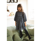 A young girl with long brown hair stands on a green couch, wearing the Louis Louise Girls’ Mahana Dress—a dark blue style with scalloped trim and long sleeves—paired with black tights. Shelves and a window appear in the background.