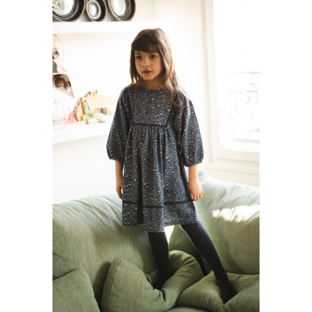 A young girl with long brown hair stands on a green couch, wearing the Louis Louise Girls’ Mahana Dress—a dark blue style with scalloped trim and long sleeves—paired with black tights. Shelves and a window appear in the background.