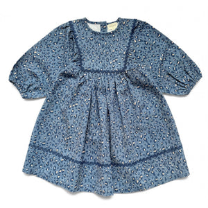 The Louis Louise Girls’ Mahana Dress is a blue long-sleeve denim dress with a loose fit, a gathered waist, decorative seam details, and an all-over leopard print pattern. It features a round neckline for added style.