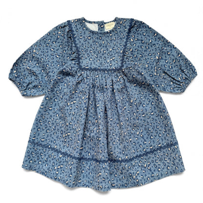 The Louis Louise Girls’ Mahana Dress is a blue long-sleeve denim dress with a loose fit, a gathered waist, decorative seam details, and an all-over leopard print pattern. It features a round neckline for added style.