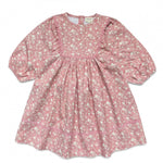 The Girls’ Mahana Dress by Louis Louise is a pink, long-sleeve floral print dress featuring white blooms, a gathered waist, and delicate ruffle details along the chest and sleeves, finished with a charming pink scalloped trim, displayed on a white background.