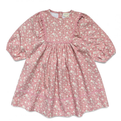 The Girls’ Mahana Dress by Louis Louise is a pink, long-sleeve floral print dress featuring white blooms, a gathered waist, and delicate ruffle details along the chest and sleeves, finished with a charming pink scalloped trim, displayed on a white background.