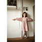 A young girl wears the Louis Louise Girls’ Mahana Dress, featuring a floral print and pink scalloped trim, as she stands against a white wall with her arms outstretched, looking at the camera.