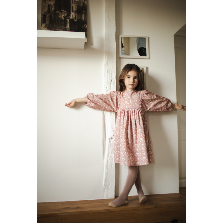 A young girl wears the Louis Louise Girls’ Mahana Dress, featuring a floral print and pink scalloped trim, as she stands against a white wall with her arms outstretched, looking at the camera.