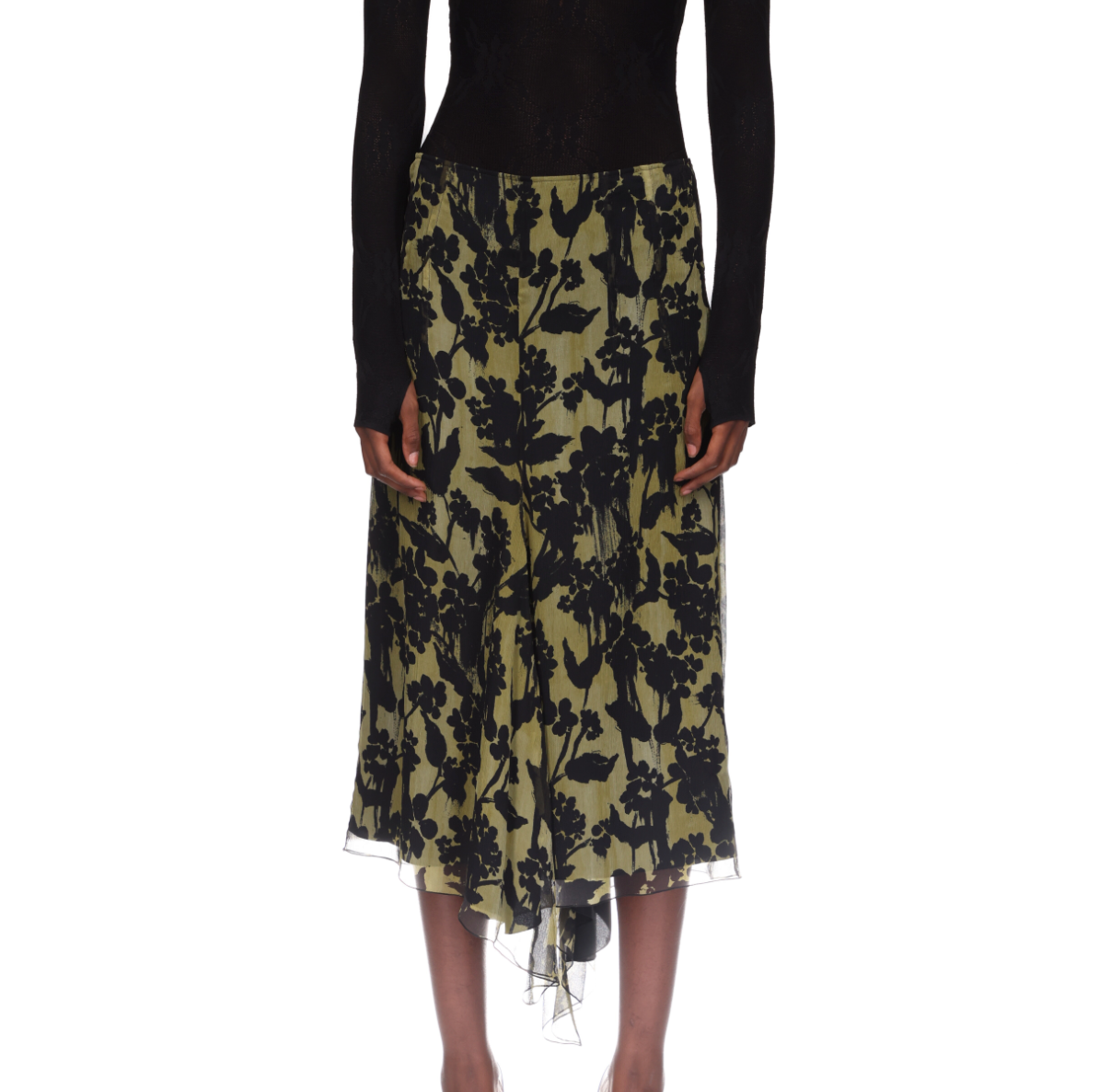 A person is seen from the front wearing a long-sleeve black top and the Victoria Beckham Corset Detail Skirt—an olive green asymmetric midi skirt with a black floral pattern.