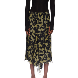 A person is seen from the front wearing a long-sleeve black top and the Victoria Beckham Corset Detail Skirt—an olive green asymmetric midi skirt with a black floral pattern.