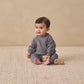 A baby wearing the Quincy Mae Baby Boys’ and Boys’ Sweatpant with an elastic waistband sits on a textured beige rug holding a wooden toy, against a plain beige background.