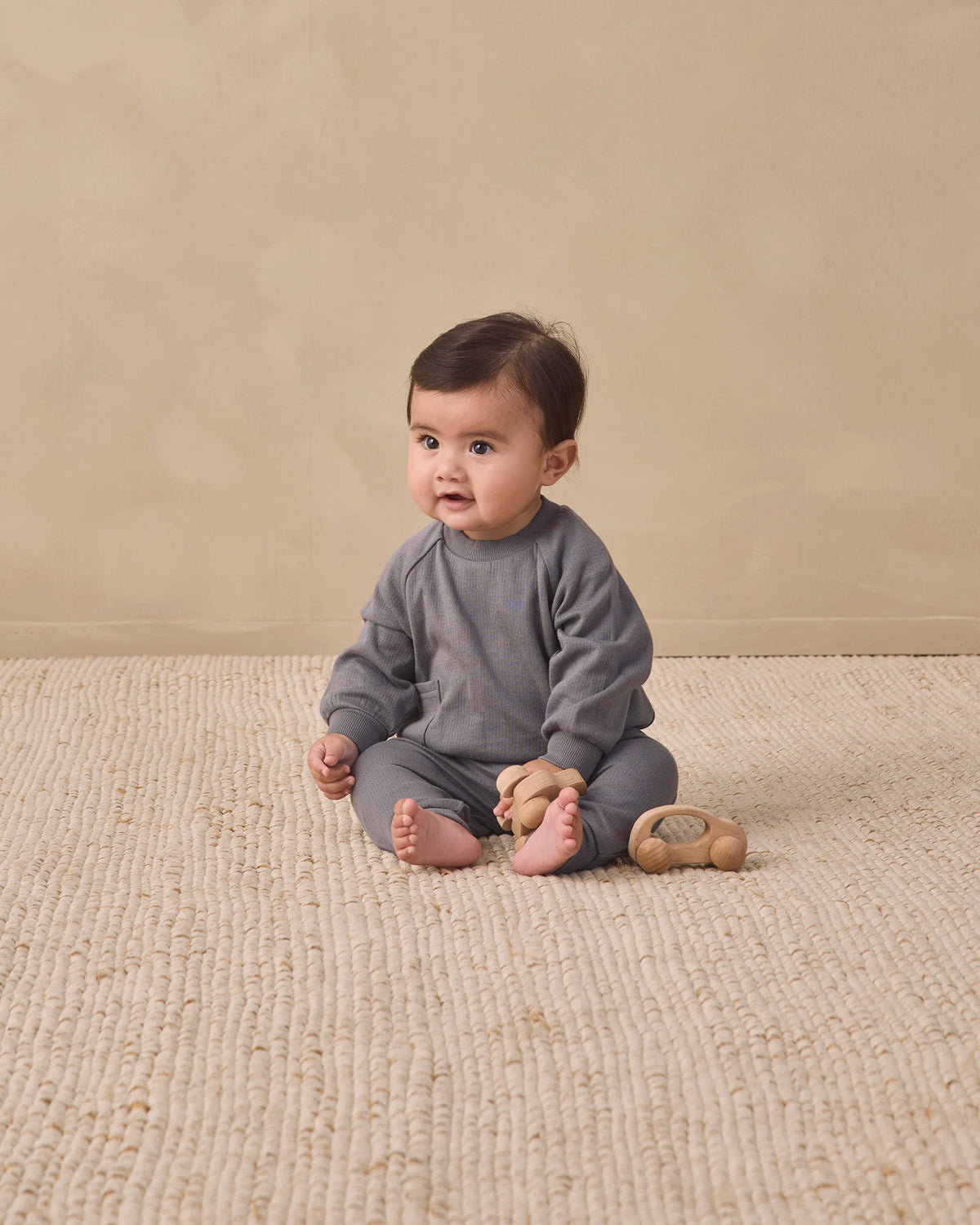 A baby wearing the Quincy Mae Baby Boys’ and Boys’ Sweatpant with an elastic waistband sits on a textured beige rug holding a wooden toy, against a plain beige background.