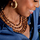 A woman wearing a blue satin shirt, Juliska Capucine De Wulf’s Earth Goddess Beads 5-Strand Necklace, and a gold hoop earring smiles while looking down.