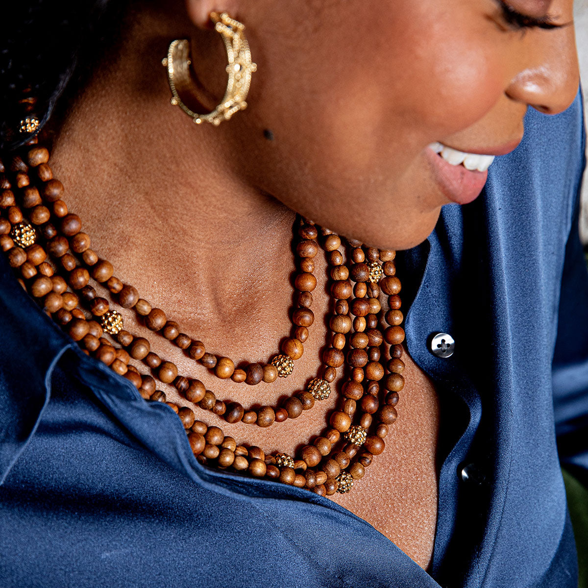 A woman wearing a blue satin shirt, Juliska Capucine De Wulf’s Earth Goddess Beads 5-Strand Necklace, and a gold hoop earring smiles while looking down.
