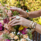 Wearing gold rings, a mother of pearl bracelet, and the Juliska Capucine De Wulf Villandry Fleur Scalloped Cuff, a person arranges a bouquet of vibrant flowers outdoors.
