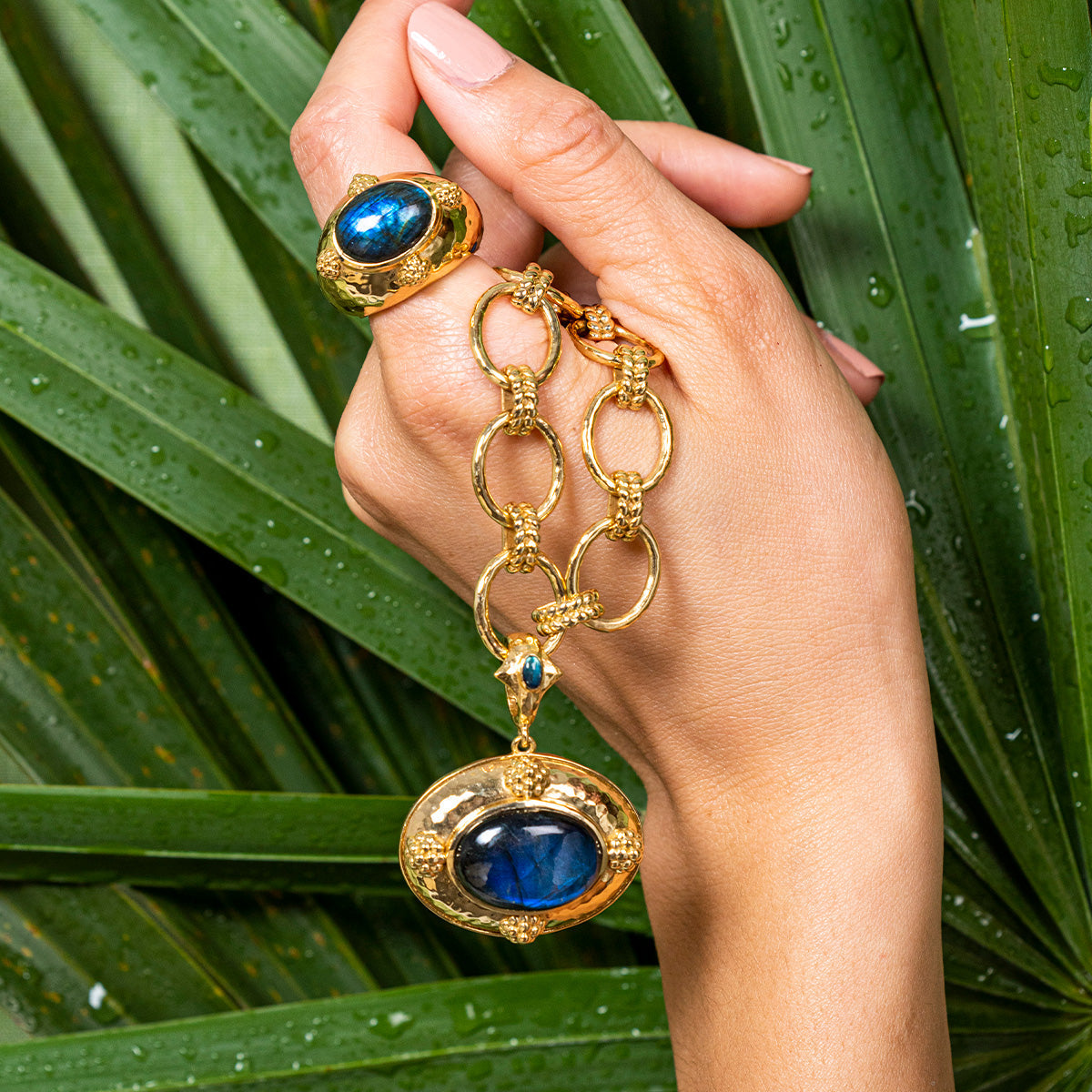 A hand displays the Juliska Capucine De Wulf Cleopatra Oval Ring in gold with blue labradorite, set against green palm leaves with water droplets.