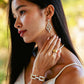 A woman with long dark hair in a white top wears Juliska Capucine De Wulf’s Pathway Post Drop Earrings, along with a bold ring and matching bracelet, as she gazes outdoors.