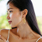 A woman with long dark hair in a white top wears Juliska Capucine De Wulf's Pathway Post Drop Earrings, gazing left with a neutral expression.