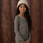 A young girl with long dark hair, wearing a cream knit hat and the Rylee & Cru Girls’ Long Sleeve Knit Tee, stands in front of a wooden wall looking slightly to the side with a neutral expression.