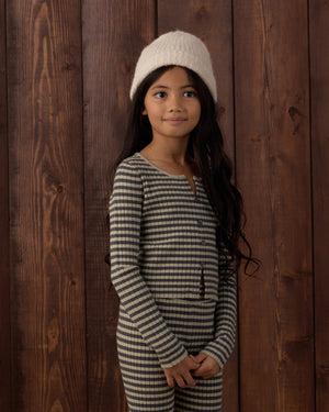 A young girl with long dark hair, wearing a cream knit hat and the Rylee & Cru Girls’ Long Sleeve Knit Tee, stands in front of a wooden wall looking slightly to the side with a neutral expression.