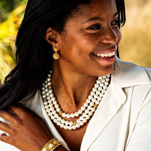 Woman wearing a white blouse, layered pearl and gold necklaces, Berry Double Drop Earrings by Juliska Capucine De Wulf, gold bracelet, smiling and looking to the side.
