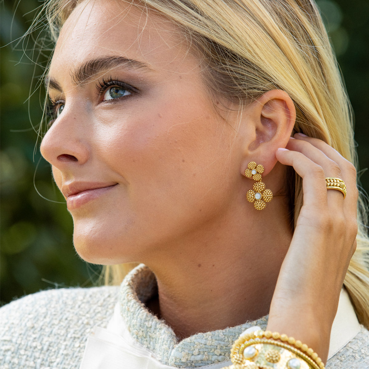 A woman wearing Juliska Capucine De Wulf Berry Clover Convertible Duo Earrings, a gold ring, and gold and pearl bracelets gazes to the side with her hand near her ear.