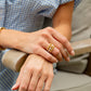 A person in a blue checkered shirt rests their hands on a wooden armrest, showcasing stacked gold rings with pearls, including the Juliska Capucine De Wulf Berry Gem Ring, on their finger.