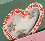 Konges Slojd Jewelery Set features cherry-shaped necklace and earrings with gold outlines, green leaves, and red cherries in a heart-shaped box window.