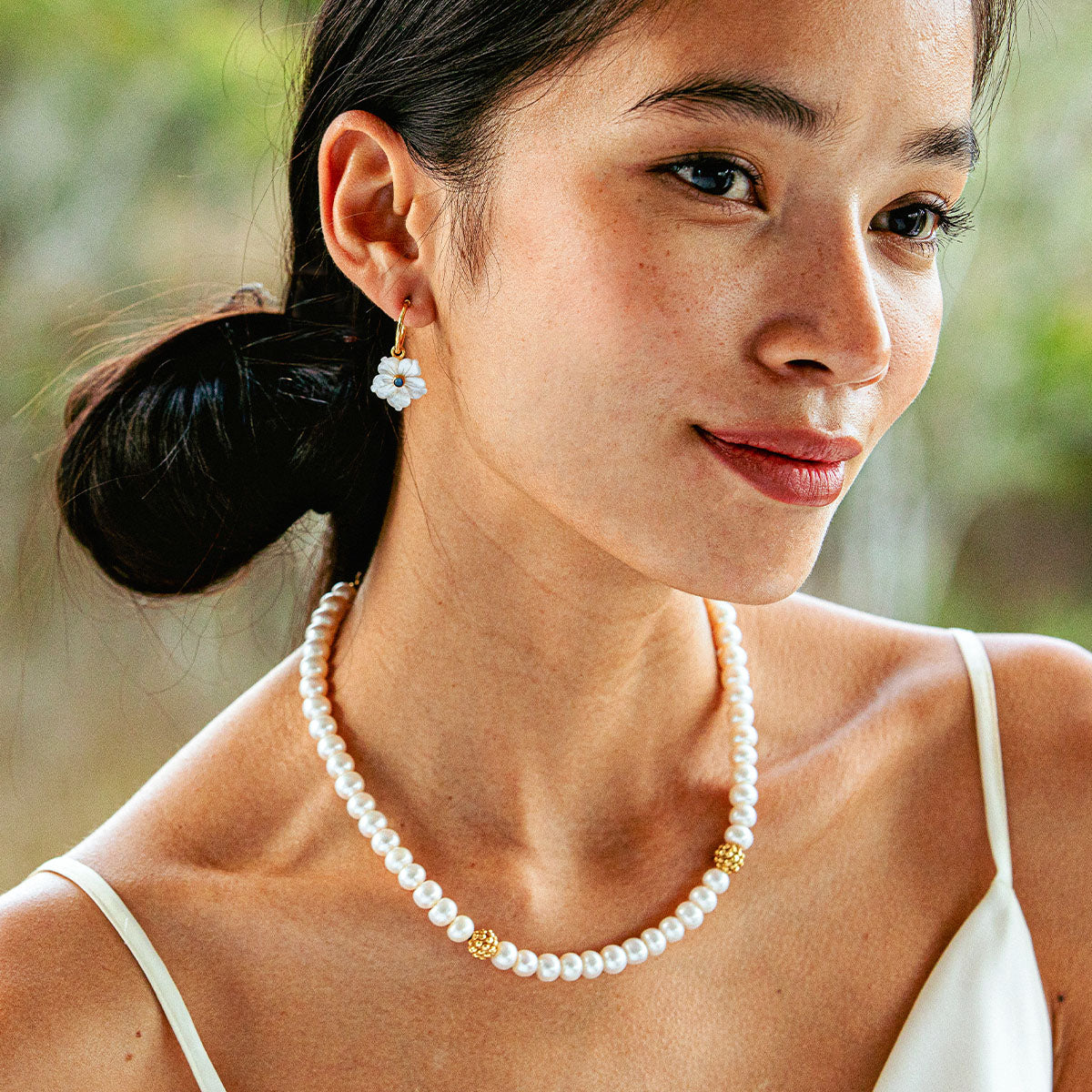 A woman wearing the Juliska Capucine De Wulf Berry Single Strand Necklace and a white flower earring, dressed in a cream spaghetti strap top, looks slightly to the side.