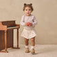 A toddler wearing the Quincy Mae Baby Girls’ Waffle Long Sleeve Bodysuit in cream stands on a textured rug, holding a red apple beside a small wooden piano set against a beige background.