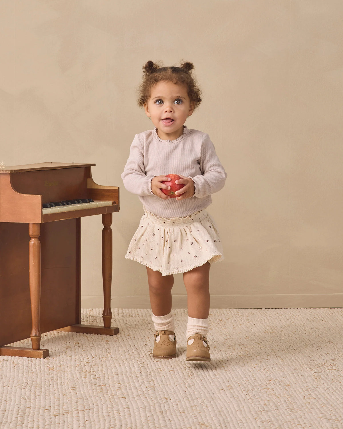 A toddler wearing the Quincy Mae Baby Girls’ Waffle Long Sleeve Bodysuit in cream stands on a textured rug, holding a red apple beside a small wooden piano set against a beige background.