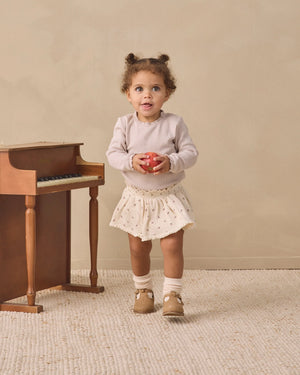 A toddler wearing the Quincy Mae Baby Girls’ Waffle Long Sleeve Bodysuit in cream stands on a textured rug, holding a red apple beside a small wooden piano set against a beige background.