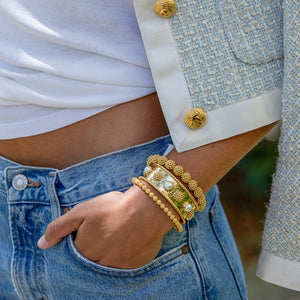 A person wearing blue jeans, a white top, a light textured jacket with gold buttons, accessorized with three gold bracelets and the Juliska Capucine De Wulf Berry Gem Cuff on their wrist.