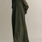 A person in TWP's Jenny's Coated Linen Gown, an olive green, oversized shirt dress, stands against a plain background with hands tucked behind their back.