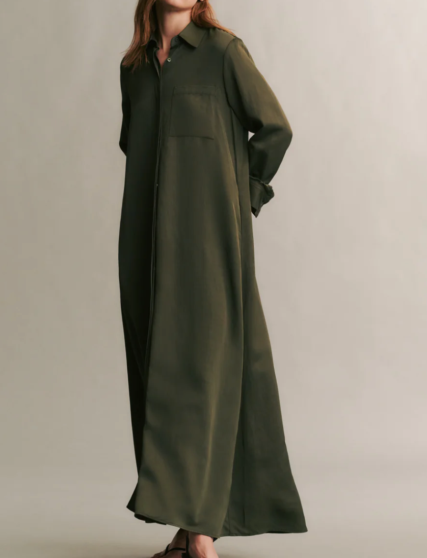 A person in TWP's Jenny's Coated Linen Gown, an olive green, oversized shirt dress, stands against a plain background with hands tucked behind their back.