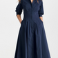 A woman stands against a plain background wearing the Simkhai Signature Jazz Dress. This navy blue, long-sleeved, A-line midi dress features pintucks and pockets. The button-up design is made from cotton poplin for style and comfort.