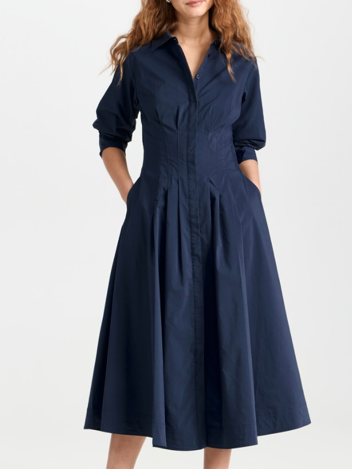 A woman stands against a plain background wearing the Simkhai Signature Jazz Dress. This navy blue, long-sleeved, A-line midi dress features pintucks and pockets. The button-up design is made from cotton poplin for style and comfort.