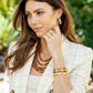 Woman in a cream plaid blazer and layered wooden bead necklaces poses outdoors, accessorized with Juliska Capucine De Wulf’s Gaia Cage Hoop Earrings, gold jewelry on her hand, wrist, and ears, and teak accents amidst lush greenery.