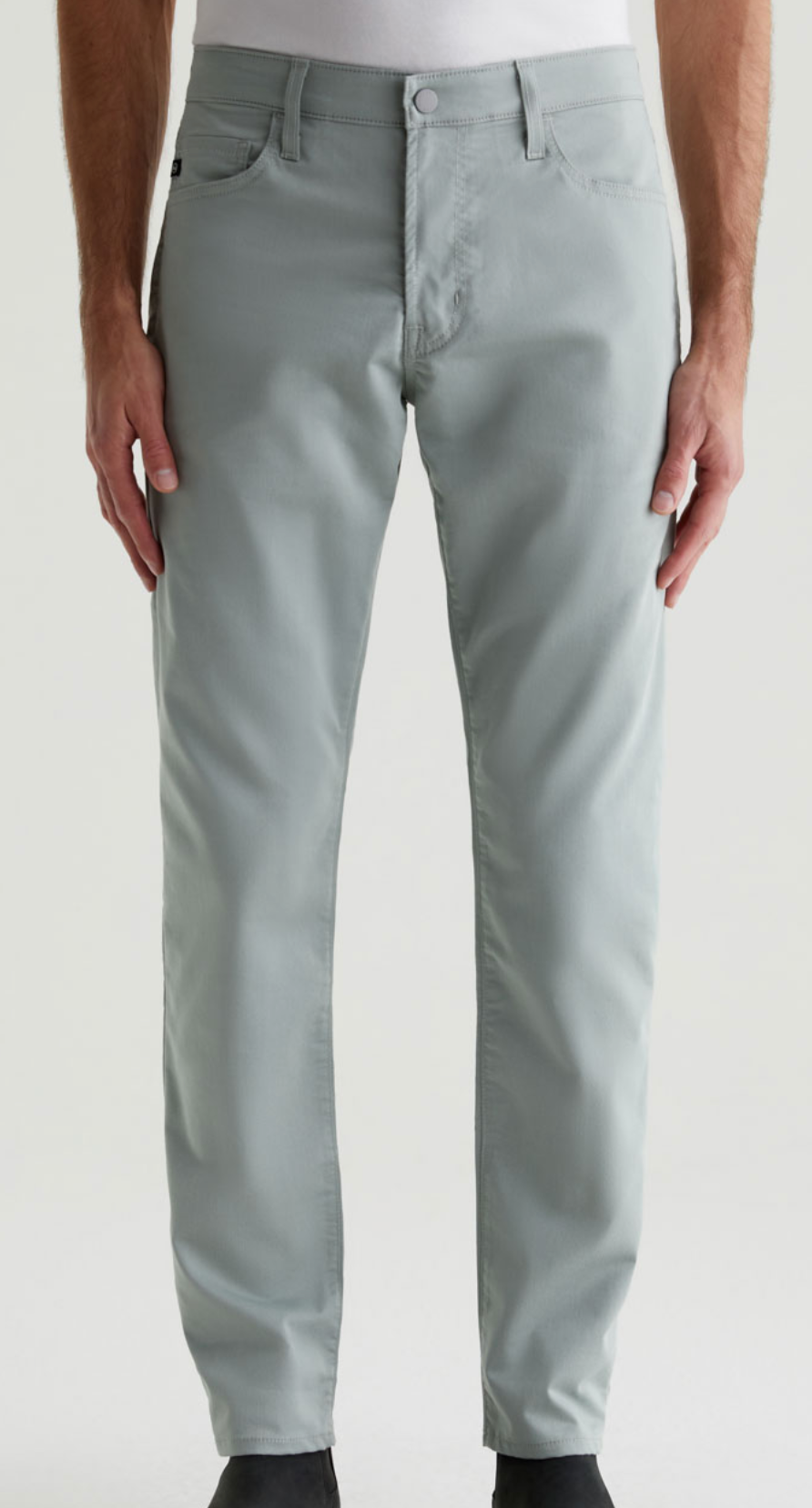 A person wearing AG Jeans Performance Airluxe™ Modern Slim men's five-pocket pants in light gray and a white shirt, standing with hands by their sides, shown from the waist down.