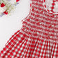 The Kidiwi Baby Girls' and Girls' Emma Smocked Dress, made of red and white gingham cotton with embroidered details, is displayed flat on a white surface beside assorted flowers.
