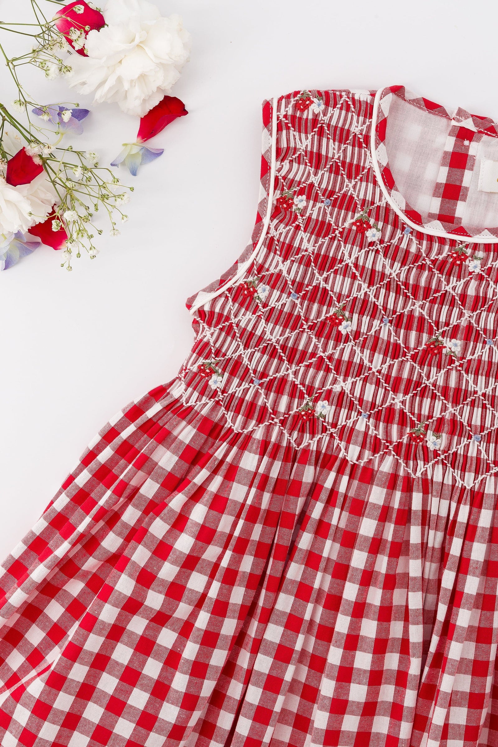 The Kidiwi Baby Girls' and Girls' Emma Smocked Dress, made of red and white gingham cotton with embroidered details, is displayed flat on a white surface beside assorted flowers.