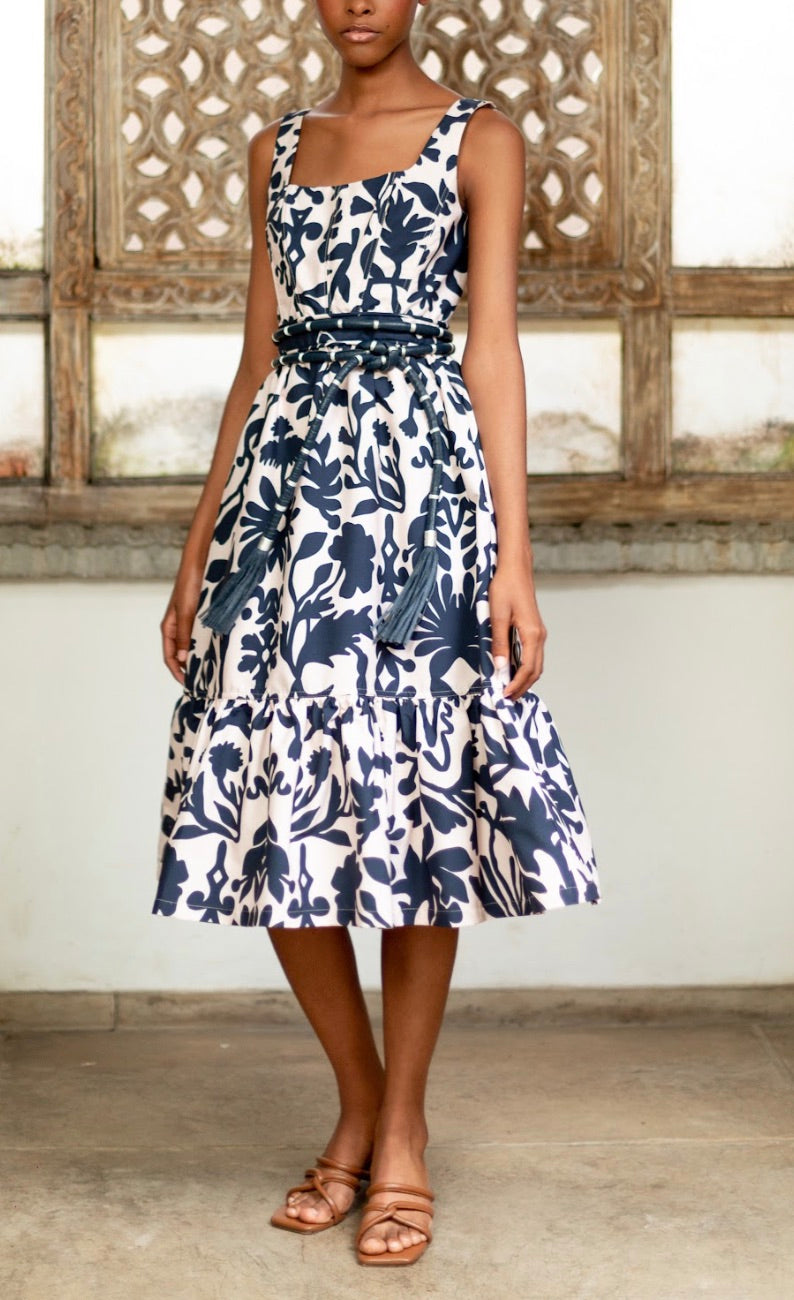 A person in an Andres Otalora Encanto Midi Dress with a classic feminine silhouette stands before a decorative wooden panel, wearing brown sandals.