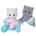 Two Calico Critters Persian Cat Twins with fluffy fur, each dressed in a lace skirt—one is seated in a blue tricycle while the other stands beside it.